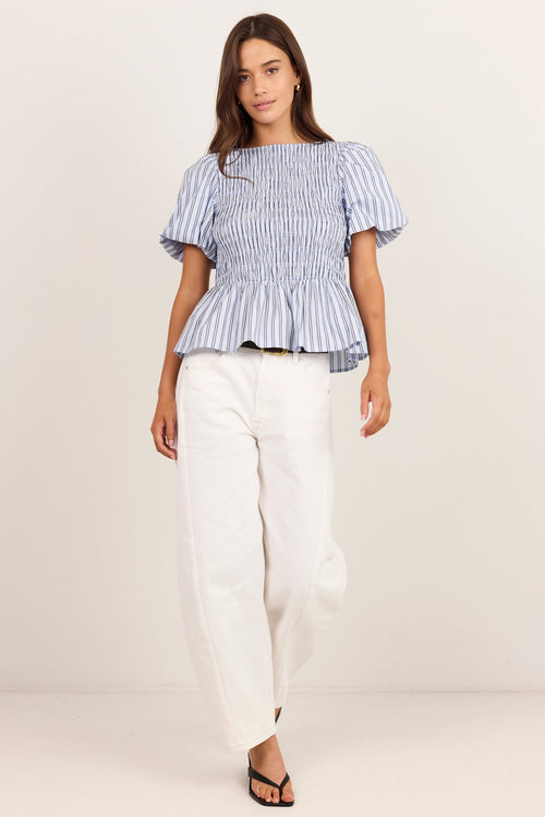 A woman modeling a blue and white vertically striped peplum top with puff sleeves, paired with white wide-leg pants and black sandals, against a plain off-white studio background.