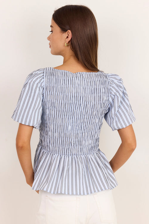 A woman shown from the back wearing a blue and white vertically striped smocked blouse with puff sleeves and a peplum hem, paired with white pants, against a plain off-white background.
