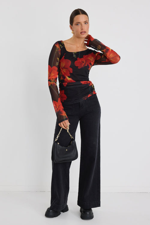 Woman wearing a fitted long sleeve mesh top with bold red floral print and sheer sleeves, paired with black jeans.