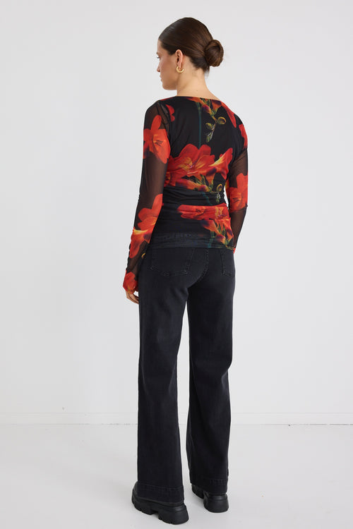 Woman wearing a fitted long sleeve mesh top with bold red floral print and sheer sleeves, paired with black jeans.