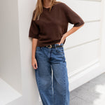 Woman wearing a brown sweater and blue jeans standing against a white wall.