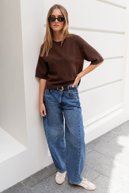 Woman wearing a brown sweater and blue jeans standing against a white wall.