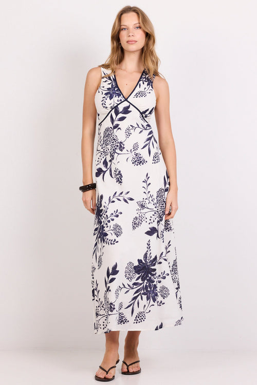 A woman modeling a sleeveless maxi dress with a navy blue floral and botanical print on a white background, standing against a plain white studio backdrop.