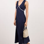 A woman models a sleeveless navy midi dress with white contrast trim and a wrap-style crossover detail at the bodice. She is holding a small beige handbag and wearing clear-strap heeled sandals against a plain white studio background.