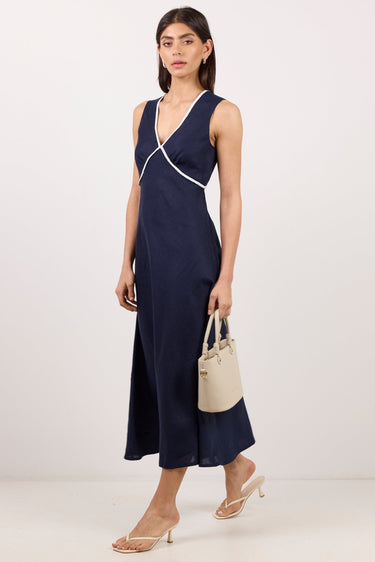 A woman models a sleeveless navy midi dress with white contrast trim and a wrap-style crossover detail at the bodice. She is holding a small beige handbag and wearing clear-strap heeled sandals against a plain white studio background.