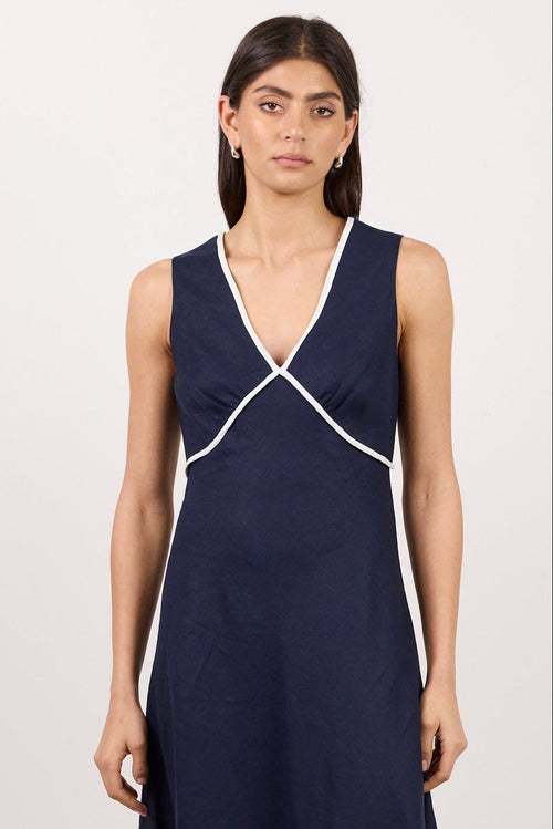 A woman modeling a sleeveless navy midi dress with white contrast piping details along the neckline and sides, standing against a plain white studio background.