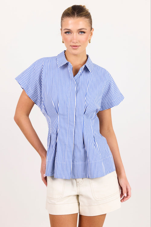 A woman modeling a blue and white vertically striped short-sleeve button-up blouse with a peplum waist, paired with cream shorts, against a plain light background.