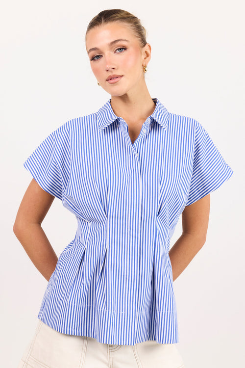 A woman modeling a blue and white vertically striped short-sleeve button-up shirt with a peplum waist, paired with cream shorts, against a plain white studio background.
