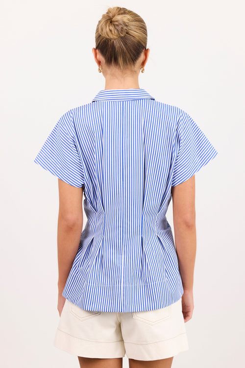 A woman viewed from the back wearing a blue and white vertically striped short-sleeve blouse with a peplum waist, paired with cream shorts, against a plain white background.