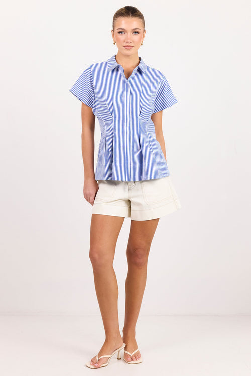 A woman modeling a light blue and white vertically striped short-sleeve button-up blouse with a peplum waist, paired with cream shorts and white heeled sandals, against a plain white studio background.