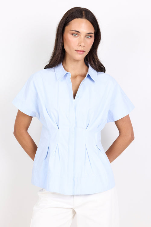A woman modeling a light blue short-sleeve button-up shirt with a peplum waist and white pants, photographed against a plain white studio background.