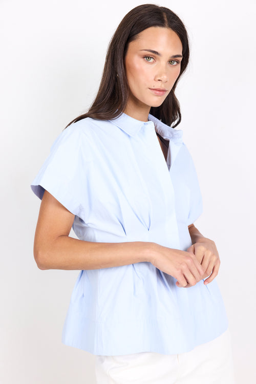 A woman modeling a light blue short-sleeve blouse with a high collar and a gathered peplum waist, paired with white pants, against a plain white studio background.