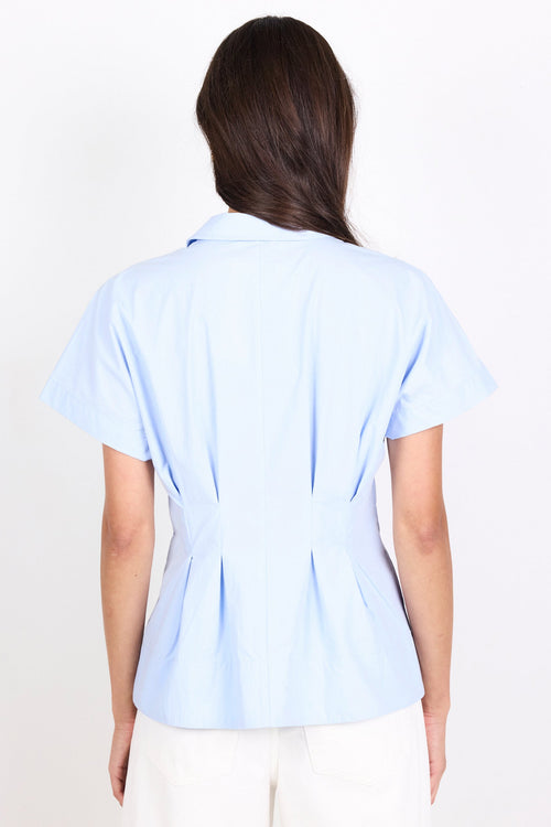 A woman viewed from the back wearing a light blue short-sleeve collared blouse with gathered detailing at the waist and white pants, against a plain white background.