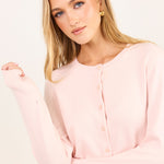 A blonde woman wearing a light pink long-sleeve button-up cardigan, posed against a clean off-white studio background.