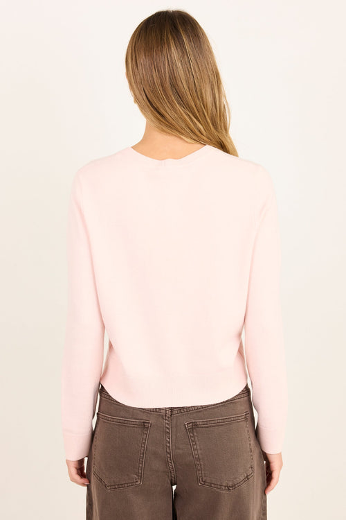 A woman viewed from behind wearing a light pink long-sleeve knit top and brown wide-leg pants against a plain off-white background.