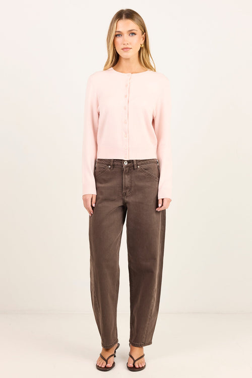 A woman stands against a plain light background wearing a pale pink long-sleeve button-front top, dark brown straight-leg jeans, and black sandals.