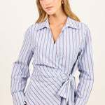 A woman modeling a light blue wrap-style button-up shirt with thin red vertical stripes and a tie waist, paired with brown pants, against a plain off-white studio background.