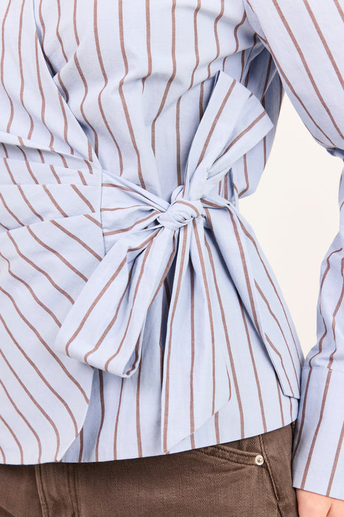A close-up of a person wearing a light blue and white vertically striped shirt with brown pinstripes, featuring a large self-tie bow at the waist, paired with brown pants.