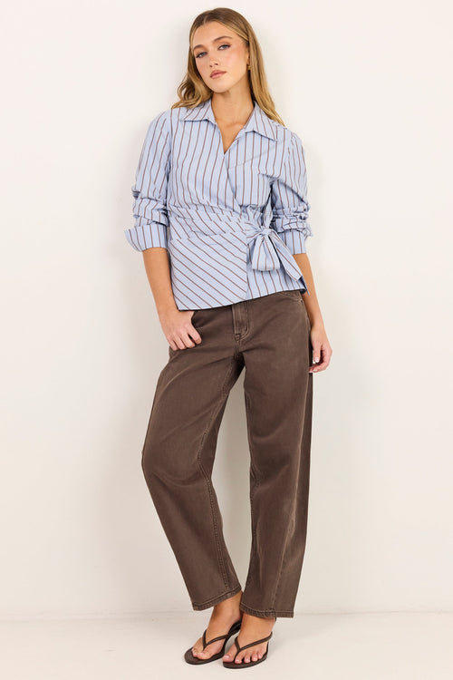 A woman modeling a light blue and white vertically striped wrap shirt with rolled sleeves, paired with brown wide-leg trousers and black sandals, standing against a plain off-white studio background.