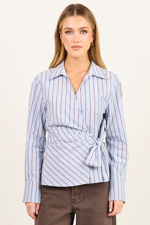 A woman stands against a plain off-white studio background wearing a light blue and white vertically striped long-sleeve wrap shirt with a tie waist, paired with brown wide-leg trousers and black heeled sandals.