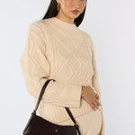 Woman wearing a beige cable knit sweater and holding a black handbag against a white background