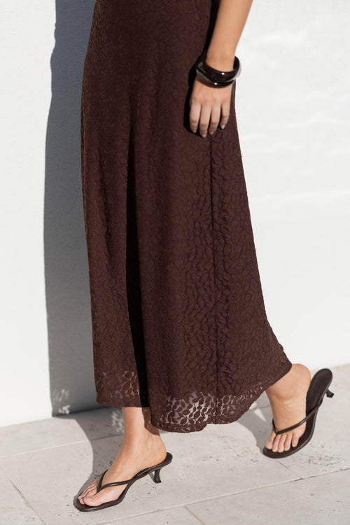 A person wearing a dark brown textured maxi skirt and black thong sandals with low kitten heels, standing on a light stone floor against a white wall.