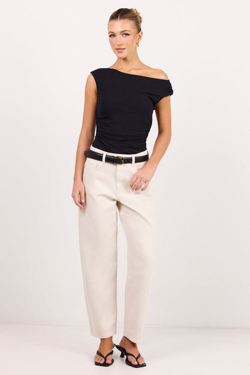 A woman modeling a black off-the-shoulder top and cream-coloured jeanswith a black belt, standing against a plain white studio background.