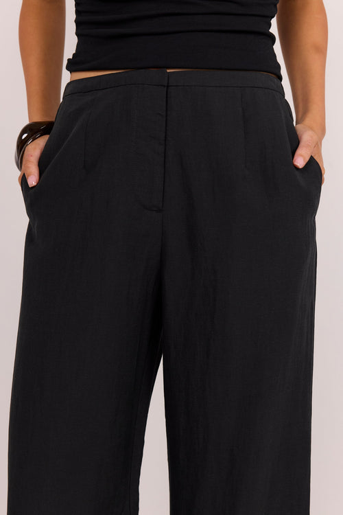 A close-up of a person wearing black wide-leg trousers with side pockets, paired with a black ribbed top, against a plain light background.
