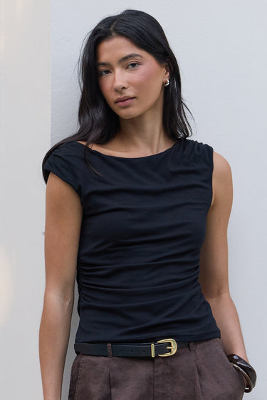 A woman wearing a black sleeveless top and brown shorts with a black belt, standing against a white building wall.