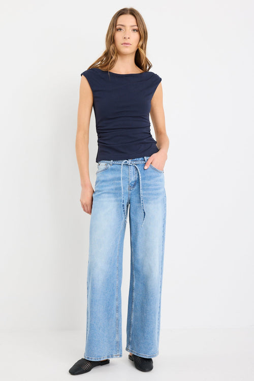 Woman wearing a fitted navy sleeveless top with boat neckline and side ruching, paired with light blue jeans.