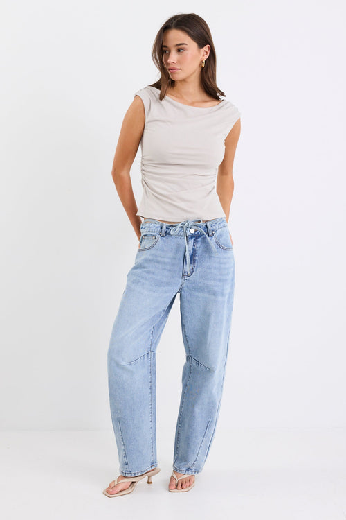 Woman wearing a stone-coloured sleeveless stretch top with a boat neckline, side ruching, and a fitted silhouette, paired with light blue jeans.