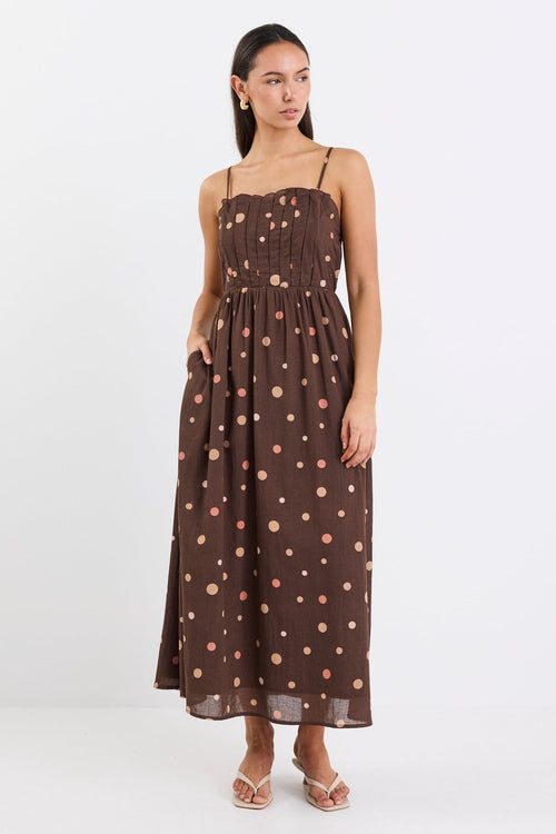 Woman wearing a chocolate brown midi dress with pink and beige polka dots, featuring a pleated bodice, thin straps, and gathered waist, styled with nude heels against a plain white background.