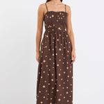 Woman wearing a chocolate brown midi dress with pink and beige polka dots, featuring a pleated bodice, thin straps, and gathered waist, styled with nude heels against a plain white background.