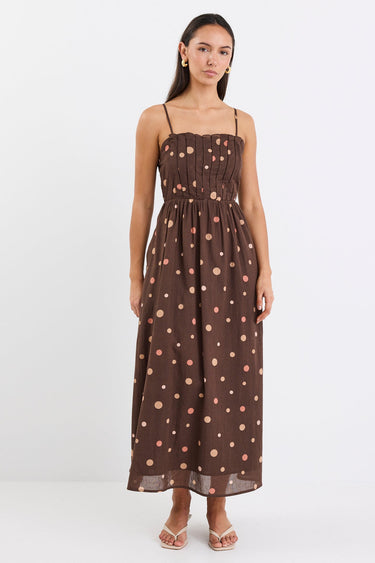 Woman wearing a chocolate brown midi dress with pink and beige polka dots, featuring a pleated bodice, thin straps, and gathered waist, styled with nude heels against a plain white background.