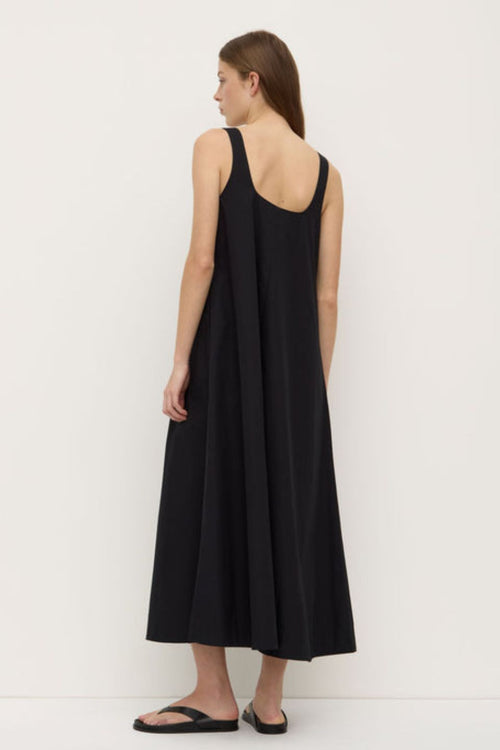 A woman wearing a black sleeveless maxi dress with wide straps and a square neckline. The dress has a flowing A-line silhouette and is styled with simple black sandals.