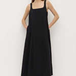 A woman wearing a black sleeveless maxi dress with wide straps and a square neckline. The dress has a flowing A-line silhouette and is styled with simple black sandals.