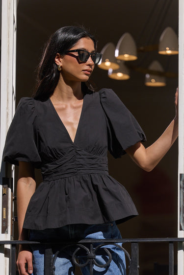 A woman wearing a black puff-sleeve peplum top and blue denim shorts stands on a balcony with white-framed doors behind her.