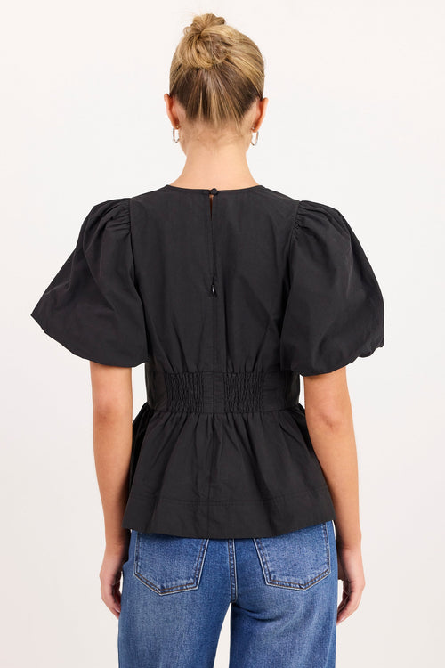 A woman viewed from the back wearing a black puff-sleeve peplum top with a smocked waist and blue jeans, against a plain white studio background.