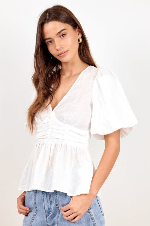 A woman modeling a white short-sleeve peplum top with puff sleeves and a ruched V-neckline, paired with light blue denim shorts against a plain white studio background.