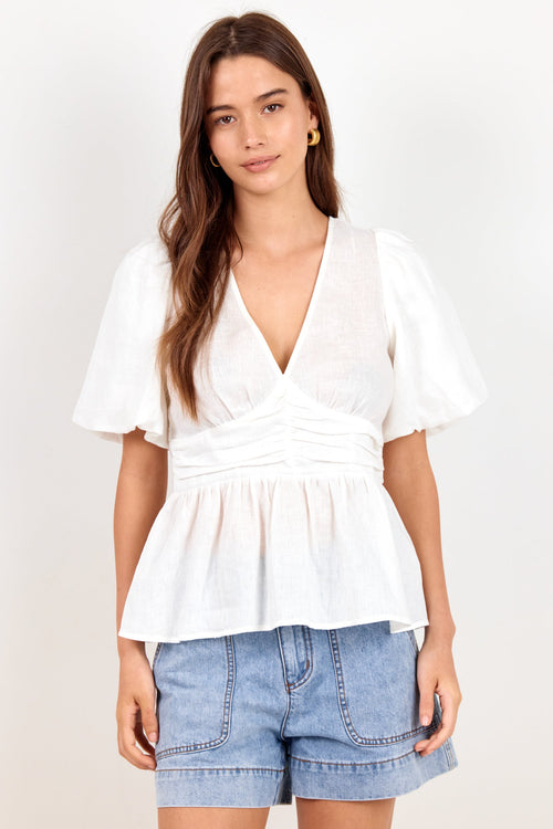 A woman modeling a white short-sleeve peplum top with a gathered waist and V-neckline, paired with light blue denim shorts, against a plain white studio background.