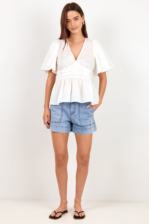 A woman modeling a white short-sleeve peplum top and light blue denim shorts against a plain white studio background.