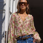 A woman wearing a beige floral long-sleeve blouse with tie-front detailing, blue jeans, and sunglasses, standing on a balcony with white architectural columns.