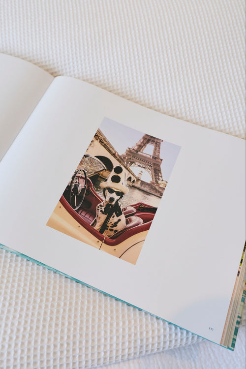 An open book or magazine lying on a white textured blanket, showing a printed photo insert with a vintage car scene and the Eiffel Tower.