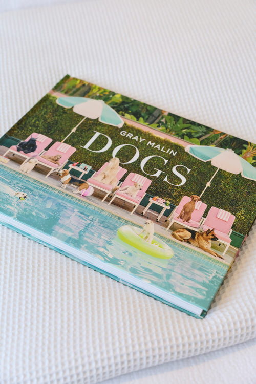The book 'Dogs' by Gray Malin, featuring a colorful cover illustration of dogs relaxing by a pool, resting on a white textured fabric surface.