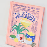 A pink hardcover book titled 'Juniper Loza' by Ellie Andrews, featuring colorful cocktail illustrations on the cover, photographed standing upright on a light marble surface against a white background.