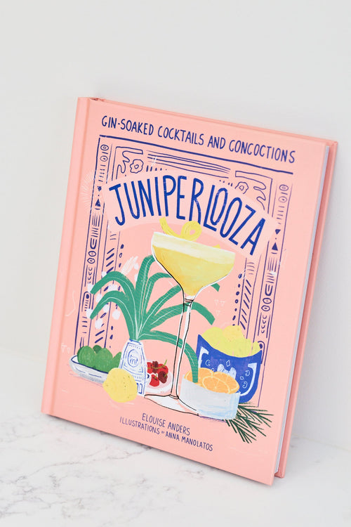 A pink hardcover book titled 'Juniper Loza' by Ellie Andrews, featuring colorful cocktail illustrations on the cover, photographed standing upright on a light marble surface against a white background.