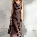 Woman in a brown evening gown holding a handbag against a light gray wall.