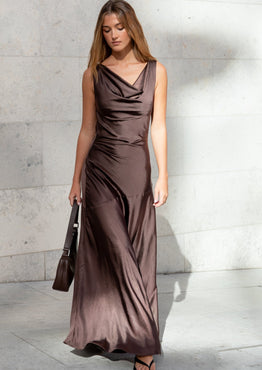 Woman in a brown evening gown holding a handbag against a light gray wall.