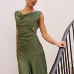 A woman wearing a sleeveless olive green maxi dress stands on an indoor staircase, holding the black metal railing. The dress has a draped, asymmetrical neckline and a flowing silhouette.