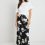 Woman wearing a black floral satin maxi skirt with a tie waist, paired with a white tee for an effortlessly chic and elegant look.